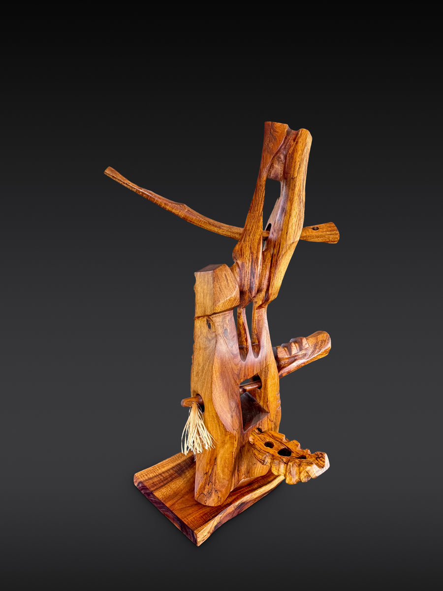 Pono, carved wood sculpture by Avi Molinas: an upright form with a long crossing arm and a fiber tassel, on a wood plinth, against a dark gray studio backdrop